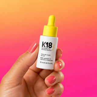 K18 Molecular Repair Hair Oil 10 mL mini with yellow dropper held between fingertips against an apricot and pink gradient.