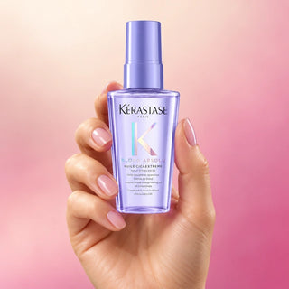 Lavender Kérastase Blond Absolu Huile Cicaextreme travel mini held in a woman's hand against a pearl and rose gradient.